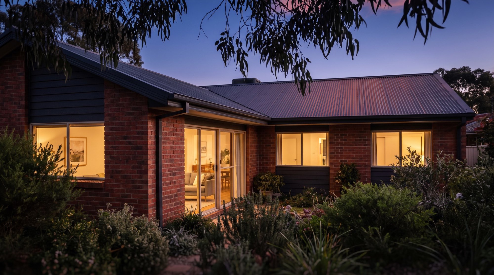 A brick home at dusk with warm light in the windows and a steel roof