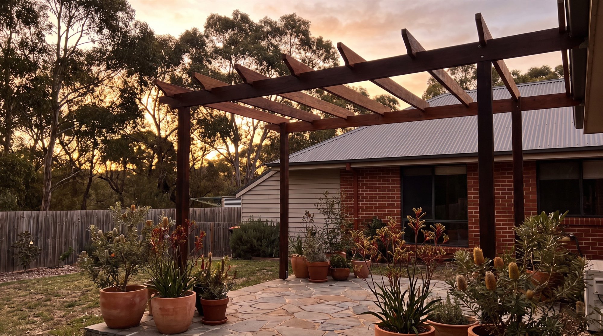 A timber pergola over a paved patio at the back of a brick home in evening light