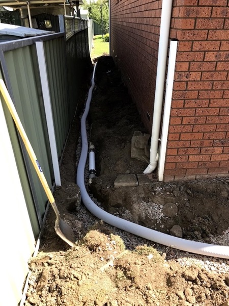 A pipe run laid into a trench beside a brick wall