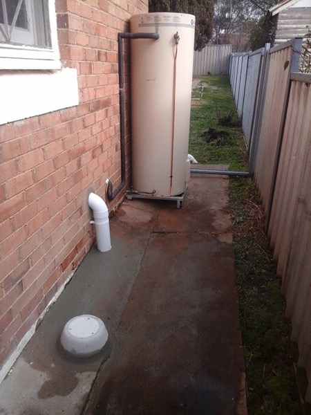 A hot water unit and downpipe on a path beside a fence