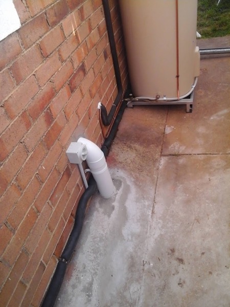 The corner of a brick wall with pipework beside a paved area